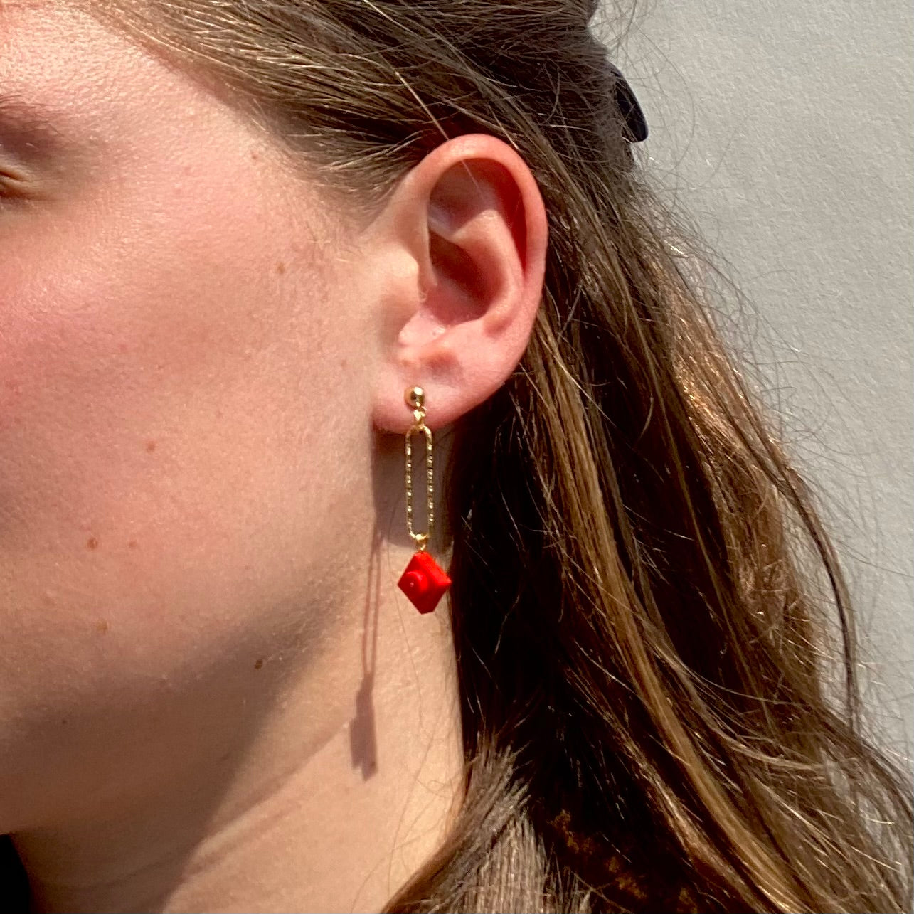 Boucle d'oreilles pendante accrochée à l'oreille par un clou d'oreille en forme de petite boule auquel est suspendu un anneau doré ovale martelé et en-dessous une brique de Lego 1x1 plate rouge vif. La boucle d'oreille est portée par une femme blanche châtain..