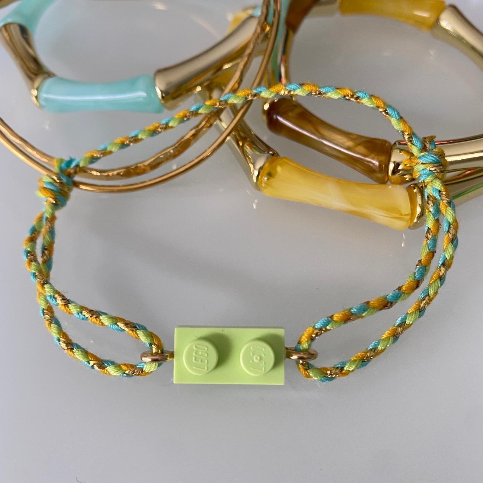 Bracelet posé à  plat avec une brique de Lego et fermé par un cordon ajustable fin tressé vert pale, orangé, turquoise et doré.  Il est présenté avec d'autres bracelets fantaisies tendance style bambou aux couleurs assorties.