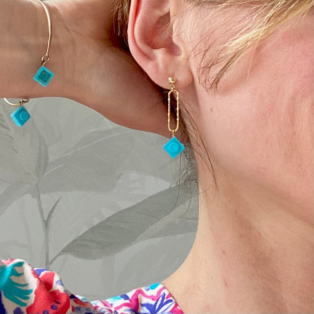 Boucle d'oreilles pendante accrochée à l'oreille par un clou d'oreille en forme de petite boule auquel est suspendu un anneau doré ovale martelé et en-dessous une brique de Lego 1x1 bleu turquoise. La boucle d'oreille est portée par une femme blanche blonde. Elle porte le bracelet Brique d'Oreille "Rockeuses" assorti.