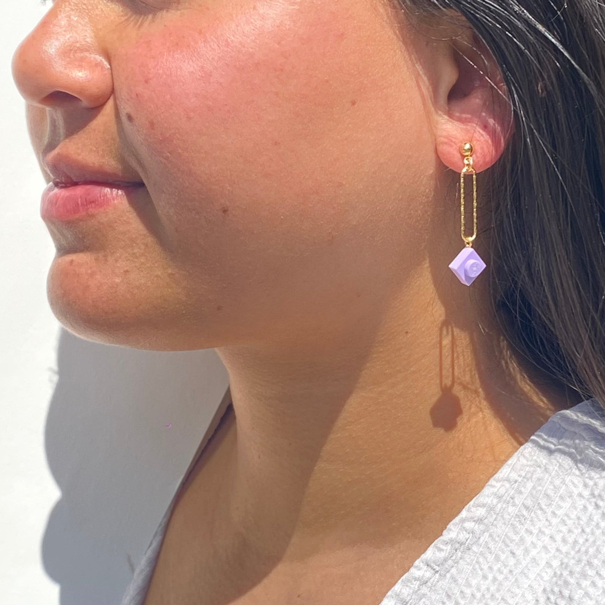 Boucle d'oreilles pendante accrochée à l'oreille par un clou d'oreille en forme de petite boule auquel est suspendu un anneau doré ovale martelé et en-dessous une brique de Lego 1x1 mauve pastel. La boucle d'oreille est portée par une femme malgache brune