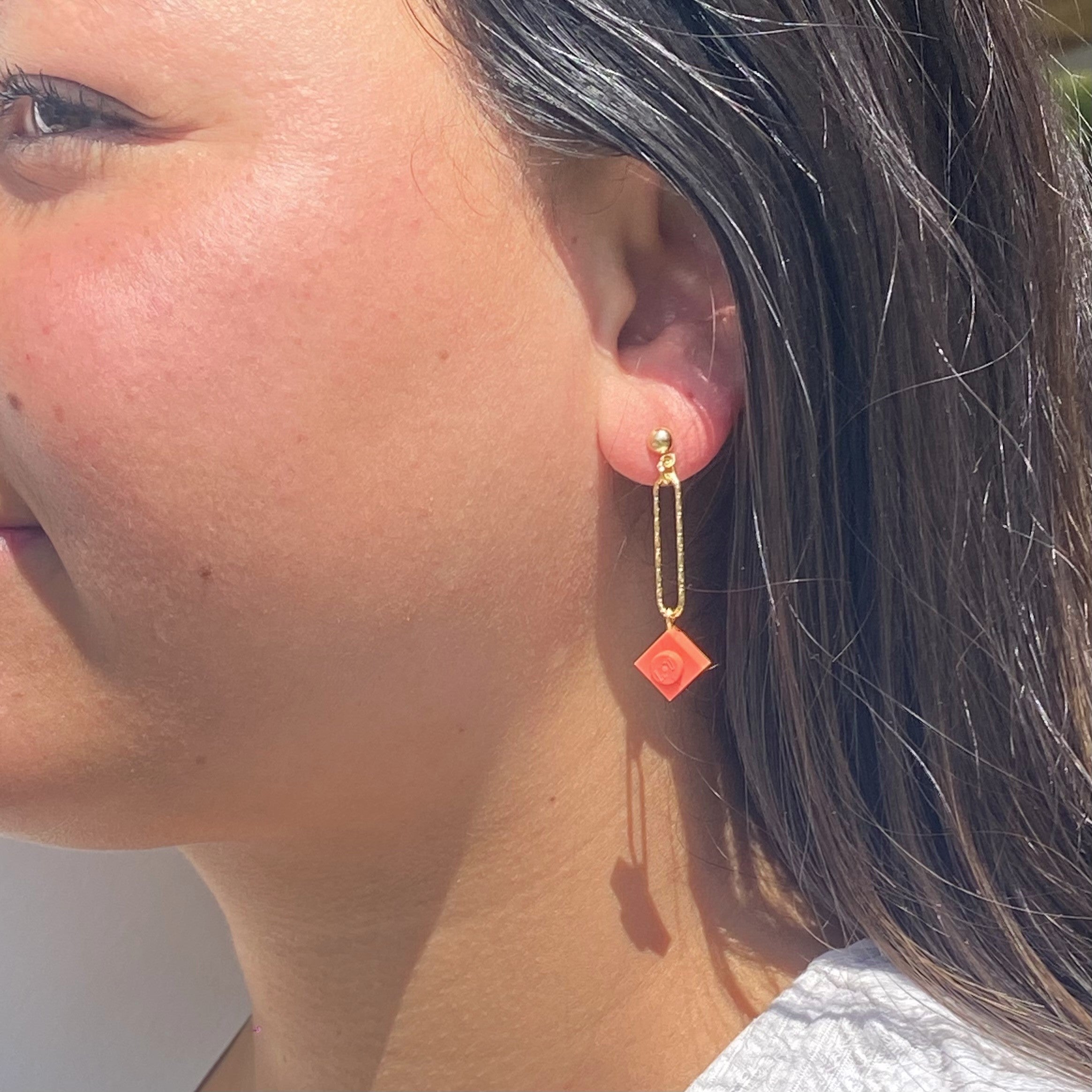 Boucle d'oreilles pendante accrochée à l'oreille par un clou d'oreille en forme de petite boule auquel est suspendu un anneau doré ovale martelé et en-dessous une brique de Lego 1x1 rose corail. La boucle d'oreille est portée par une femme malgache brune 