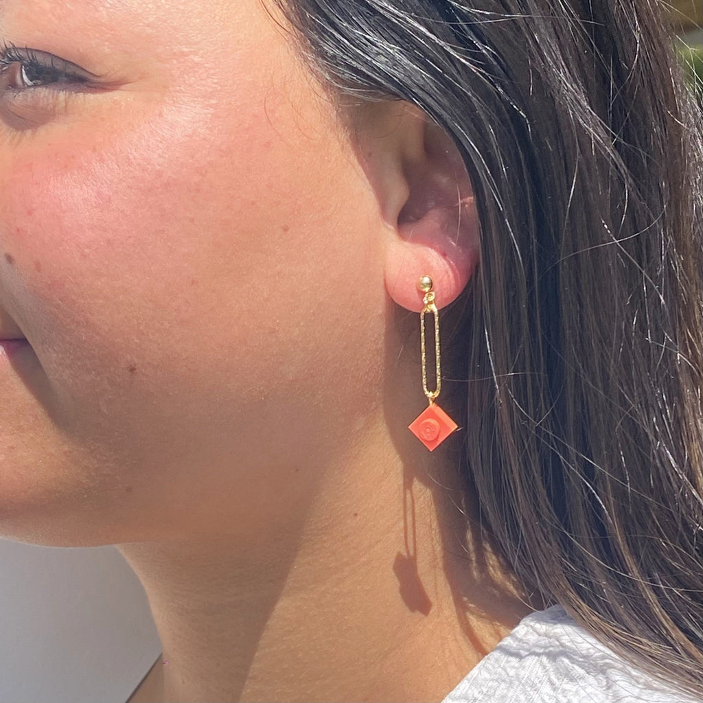 Boucle d'oreilles pendante accrochée à l'oreille par un clou d'oreille en forme de petite boule auquel est suspendu un anneau doré ovale martelé et en-dessous une brique de Lego 1x1 rose corail. La boucle d'oreille est portée par une femme malgache brune 